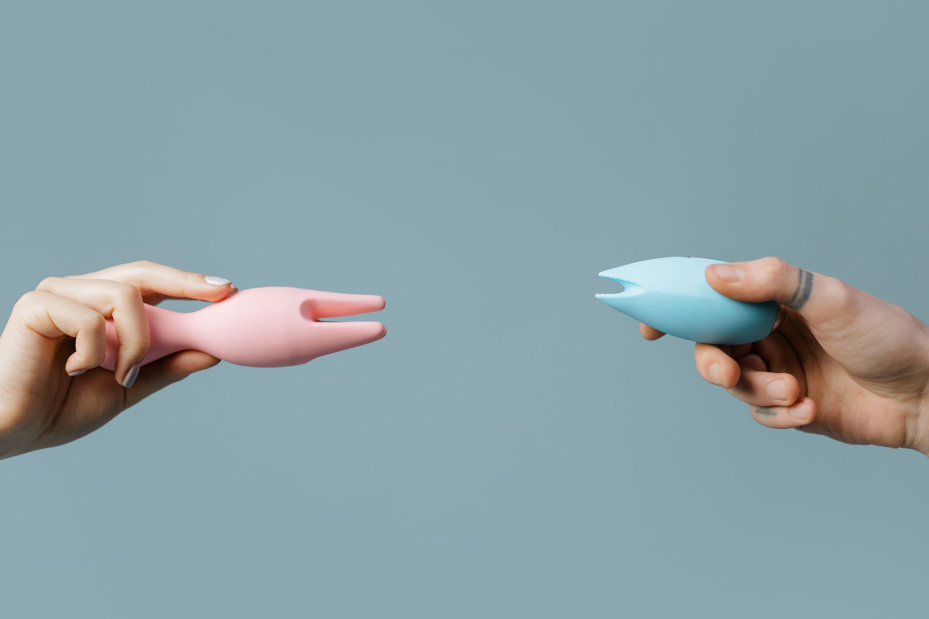 Deux mains tenant des vibrateurs en silicone rose et bleu contre un fond pastel