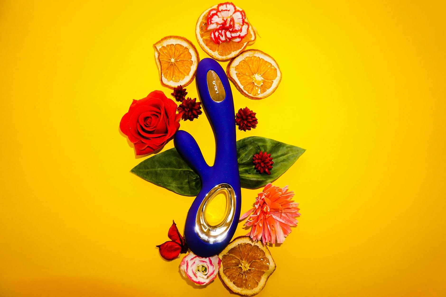 A blue vibrator surrounded by flowers and citrus on a bright yellow background, symbolizing renewal and pleasure.